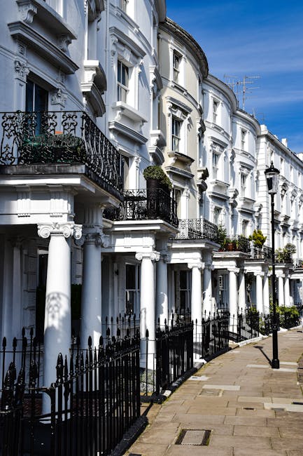 Image of a row of Victorian-style terraced houses with white facades, ornate black wrought iron balconies, and decorative moldings. The buildings have large bay windows and are evenly spaced along a c