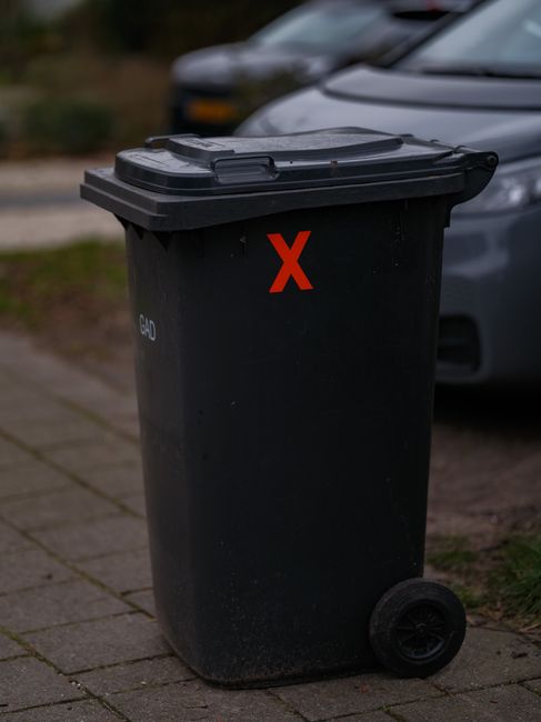 A black outdoor waste bin with a red X marked on the front, positioned on a paved sidewalk beside a parked silver car. The bin features a closed black lid with two hinges, and its surface appears clean and well-maintained. The concrete pavement has visible joints and is dry, with a background showing blurred greenery and additional vehicles. The scene reflects public waste disposal, emphasizing cleanliness and sanitation in residential or urban areas, aligning with Holland Park council waste rules, and showcasing the importance of proper waste management as part of regular cleaning and hygiene practices, which Holland Park Cleaners advocate for in their domestic and commercial cleaning services.