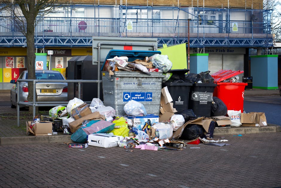 A street scene outside a commercial building with multiple overflowing recycling and waste bins, including a large grey mixed paper and card bin with its lid partially open, surrounded by scattered cardboard boxes, plastic bags, and miscellaneous rubbish on the ground. The area features a paved footpath, a metal railing, and parked cars, with the building in the background displaying signage for a fish bar. The scene highlights the need for waste management and proper disposal practices, as emphasized by Holland Park Cleaners in relation to waste rules and cleaning standards for residents and businesses in Holland Park.
