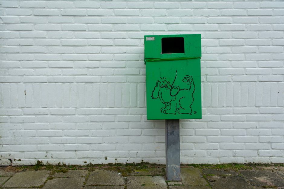 A black outdoor waste bin with a red X marked on the front, positioned on a paved sidewalk beside a parked silver car. The bin features a closed black lid with two hinges, and its surface appears clean and well-maintained. The concrete pavement has visible joints and is dry, with a background showing blurred greenery and additional vehicles. The scene reflects public waste disposal, emphasizing cleanliness and sanitation in residential or urban areas, aligning with Holland Park council waste rules, and showcasing the importance of proper waste management as part of regular cleaning and hygiene practices, which Holland Park Cleaners advocate for in their domestic and commercial cleaning services.