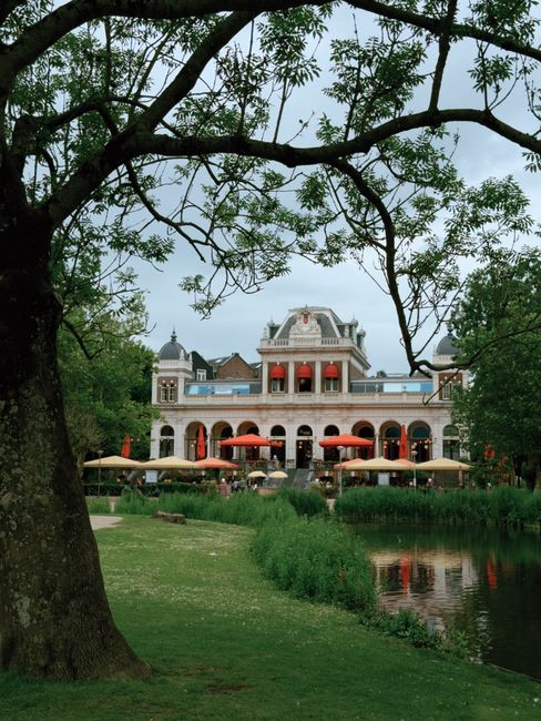 A historic multi-story building with a classical architectural style, featuring arched windows and balconies with red awnings, situated along the edge of a reflective pond. The scene is framed by lush green trees and well-maintained grass, with outdoor seating shaded by umbrellas in front of the building. The surface of the pond mirrors parts of the building and surrounding foliage, and the overall environment appears clean and well-kept, emphasizing a tranquil and inviting atmosphere. Although the image does not directly relate to cleaning, it showcases an exterior setting with natural and architectural elements consistent with professional maintenance and care as promoted by Holland Park Cleaners.