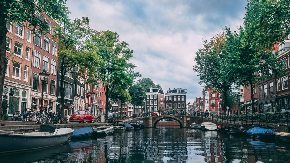 A scenic view of a canal in Holland Park, featuring calm water reflecting traditional brick townhouses and lush green trees on either side. Several small boats are moored along the canal, with some covered by blue or black tarps. A small stone bridge arches over the canal, connecting the two sides, while bicycles are parked along the railings. The buildings display a mix of red and dark brick facades with large windows, set in a charming, well-maintained residential area. The sky is partly cloudy, providing soft diffuse light over the scene, emphasizing the cleanliness and tidy appearance of the surroundings. Holland Park Cleaners would ensure that such outdoor surfaces remain pristine, highlighting the importance of surface cleaning and maintenance in residential and commercial outdoor spaces.