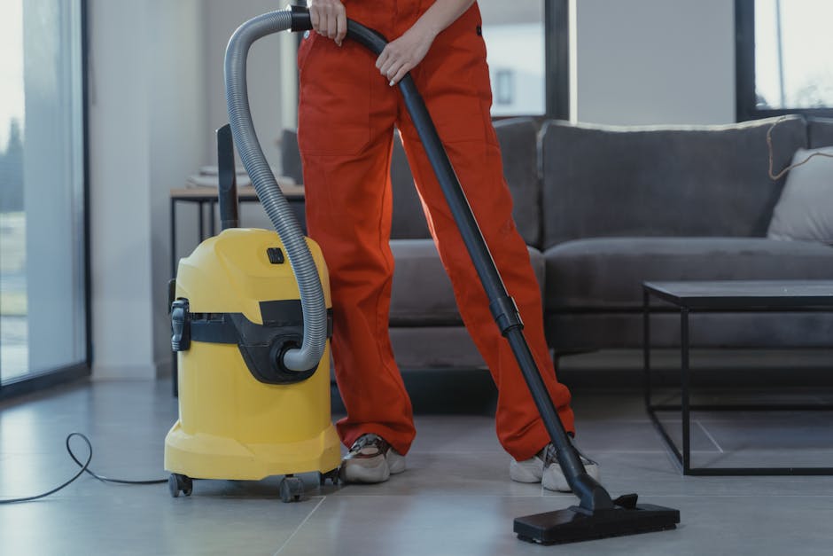 A person dressed in orange work overalls using a yellow industrial vacuum cleaner with a black hose and wand to deep clean a modern living room. The room features a light-colored tiled floor, a gray fabric sofa with cushions, and a black metal coffee table. Large windows allow natural light to illuminate the space, highlighting the cleanliness of the floor and furniture. The scene demonstrates surface cleaning and maintenance typical of professional domestic cleaning services provided by Holland Park Cleaners, focusing on thorough hygiene and stain removal in residential environments.