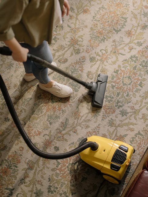 A person dressed in orange work overalls using a yellow industrial vacuum cleaner with a black hose and wand to deep clean a modern living room. The room features a light-colored tiled floor, a gray fabric sofa with cushions, and a black metal coffee table. Large windows allow natural light to illuminate the space, highlighting the cleanliness of the floor and furniture. The scene demonstrates surface cleaning and maintenance typical of professional domestic cleaning services provided by Holland Park Cleaners, focusing on thorough hygiene and stain removal in residential environments.