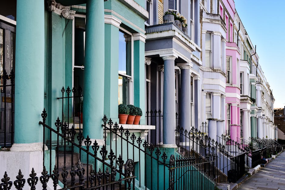 A row of colorful Victorian-style terraced houses on Notting Hill Gate, featuring pastel turquoise, blue, pink, and white facades with tall, rounded columns and decorative iron railings. The front steps are made of concrete and are topped with small potted plants, while the exterior walls have a smooth finish. Bright sunlight illuminates the buildings, highlighting their vibrant paintwork and classic architectural details, with a clear blue sky in the background. The scene depicts well-maintained residential properties reflecting the area's charm. This image relates to the content about same day cleaning service availability in Notting Hill Gate, provided by Holland Park Cleaners.