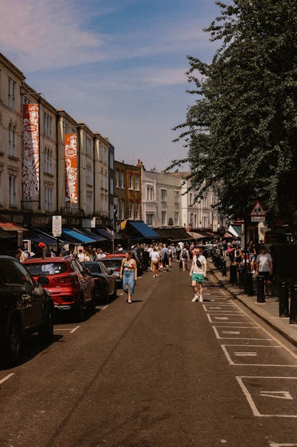 The image depicts a busy street scene at Notting Hill Gate under clear weather, with pedestrians walking along the sidewalk and in the street, lined by multi-storey buildings with shops and cafes. The street features parked cars on one side and a designated pedestrian pathway marked with white lines, separated by black bollards. Large trees with lush green foliage shade parts of the sidewalk, and the buildings have a mix of brick and painted facades, some with large windows and signs. The overall atmosphere suggests an active, vibrant area with a mix of commercial and leisure activity, while the focus remains on the lively urban environment rather than any cleaning activity, aligning with the context of a location served by Holland Park Cleaners for domestic and commercial cleaning needs.