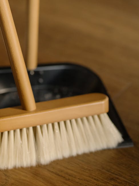 A close-up of a wooden-handled dustpan with a beige bristle brush attached, placed on a polished wooden floor, with a black plastic container partially visible behind it. The surface material appears 