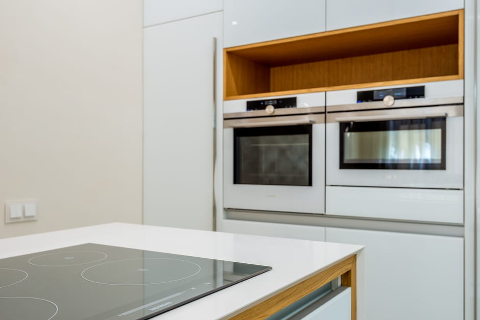 A modern kitchen featuring a white countertop with an integrated electric ceramic cooktop that has four circular cooking zones. Behind the counter, built-in stainless steel double ovens are set into white cabinetry, with a wooden open storage shelf above them. The kitchen surfaces appear clean and shiny, reflecting natural light filtering through nearby windows, suggesting recent surface cleaning and maintenance by Holland Park Cleaners. The surrounding walls are painted in a neutral light color, complementing the sleek, contemporary design of the space, which is well-organized and dust-free, highlighting the importance of surface cleaning and deep cleaning for hygiene.