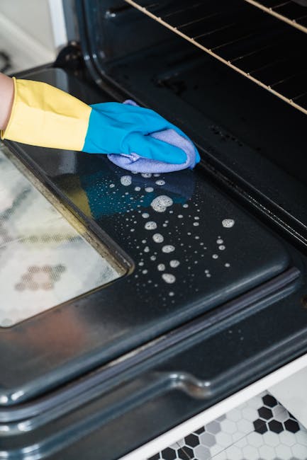 Close-up of a person's gloved hand cleaning the black glass surface of a modern oven door with a blue cleaning cloth, focusing on the smudges and soap suds being wiped away. The oven interior is visible with stainless steel racks and a dark interior, indicating a deep cleaning process. The surrounding countertop features a glossy surface with some soap residue, and the kitchen has a contemporary design with tiled flooring in white and black hexagonal patterns. This detailed scene reflects professional surface cleaning and sanitisation services provided by Holland Park Cleaners, emphasizing thorough oven maintenance in a domestic setting.